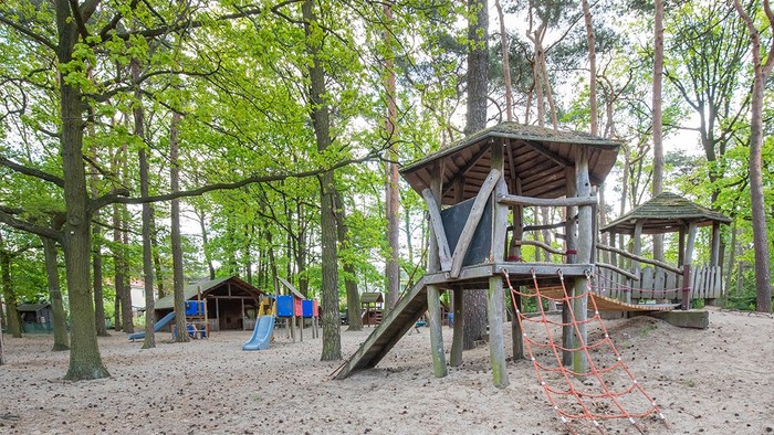 Spielplatz in einem bewaldeten Bereich mit einer hölzernen Spielanlage, Rutsche und Sandkasten.