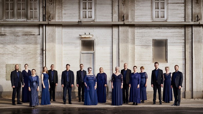 En gruppe af mænd og kvinder i formelle blå tøjer står foran et stort, koldt og gråt stenhus med flere vinduer.