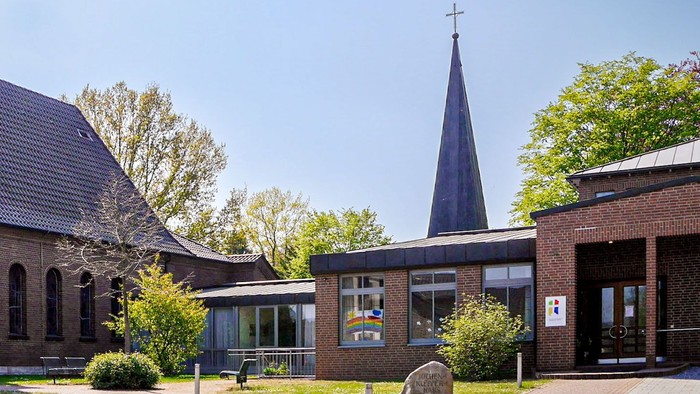 Eine Backsteinkirche mit einem spitzen Dachreiter, daran rechts angebaut ein flacher Bau vor einem Glockenturm