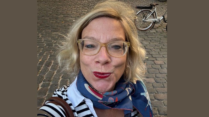 Eine blonde Frau mit Brille posiert im Freien auf einer Kopfsteinpflasterstraße.