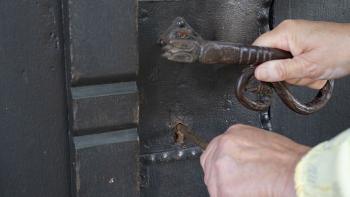 Nahansicht, wie eine rechte Hand eine Klinke drückt, während die linke Hand einen Schlüssel in das Schlüsselloch schiebt.