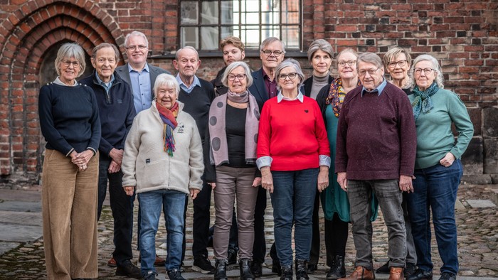 En gruppe af 16 personer står sammen foran en mur af røde mursten.