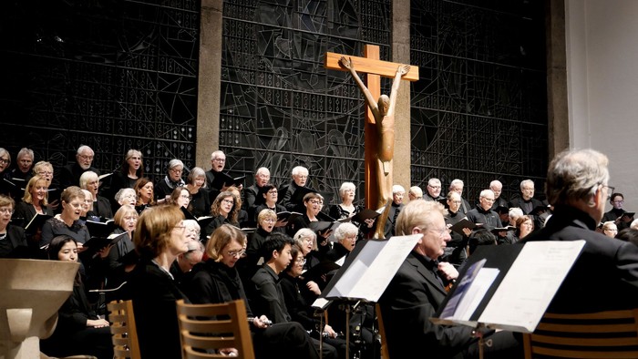 Ein Chor singt in einer Kirche vor einem dunklen Kirchenfenster und modernen Kruzifix, gesehen über die Schultern eines davor sitzenden Orchesters. Alle tragen Schwarz