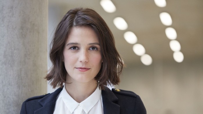 Eine junge Frau mit dunklen Haaren, einer hellen Bluse und einem marineblauen Blazer lehnt an eine Betonsäule. Im Hintergrund eine hell ausgeleuchtete Halle. 