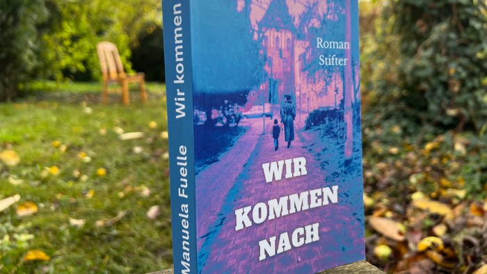 Das Buch steht auf einer Bank, dahinter ein Stuhl und ein Baum.