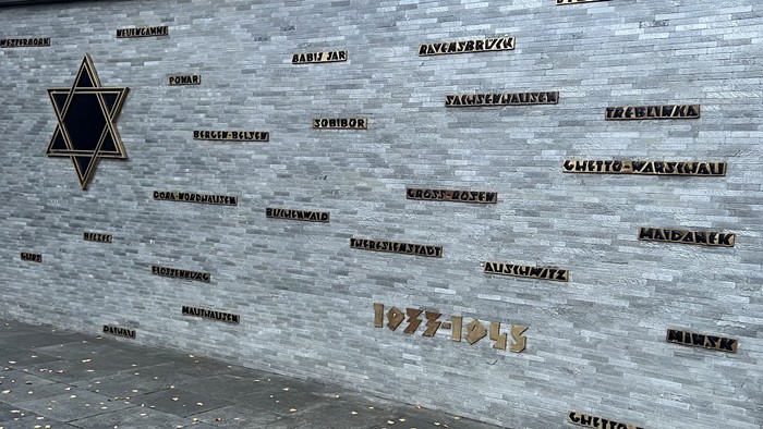 Eine graue Gedenkwand, darauf stelen die Namen der Konzentrationslager und Ghettos, in die Jüdinnen und Juden deportiert wurden von 1933-45.