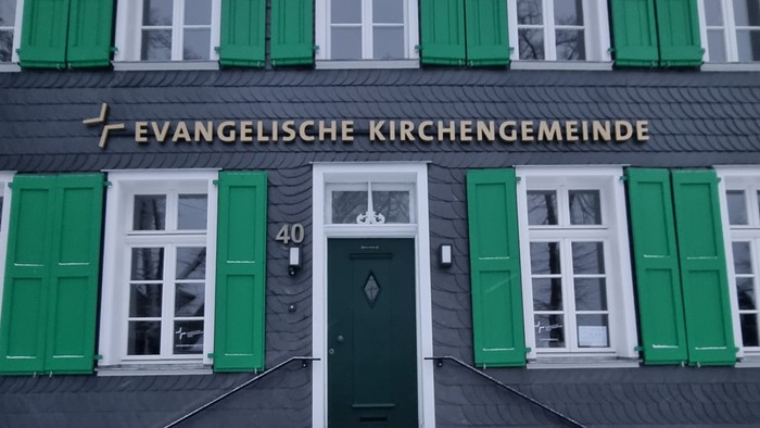 Das Bild zeigt ein Gebäude mit grünen Fensterläden und einer Treppe zum Eingang.