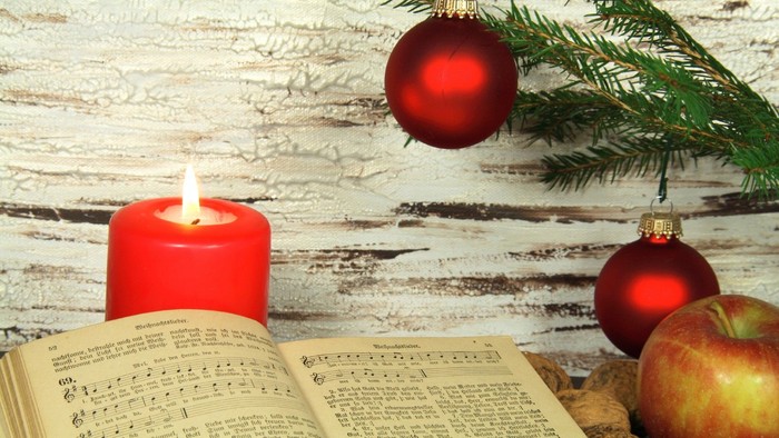 Offenes Buch mit Notenblättern, rote Kerze, rote Dekorationen und Äpfel auf einer rustikalen Holzoberfläche.