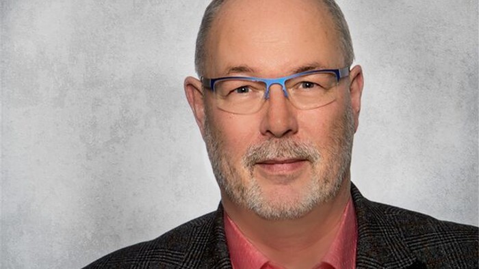 Mann mittleren Alters mit Brille und Bart, der ein Sakko und ein rosa Hemd trägt.