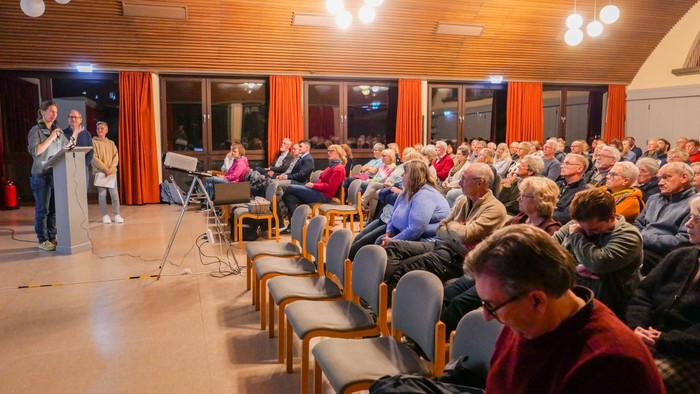Drei Vortragende stehen links vor einem vollbesetzten Saal