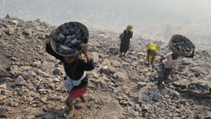 Drei Personen tragen große Bündel auf dem Rücken, während sie über ein felsiges Gelände laufen.