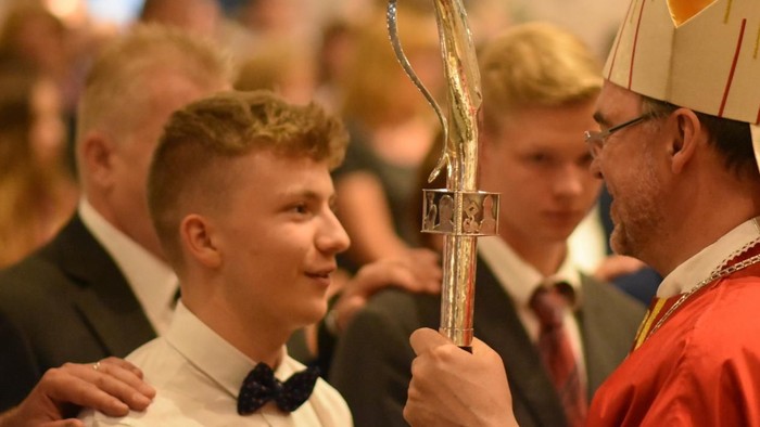 Ein Mann in einem roten Gewand und hohem Hut überreicht einem jungen Mann im Smoking einen Pokal.