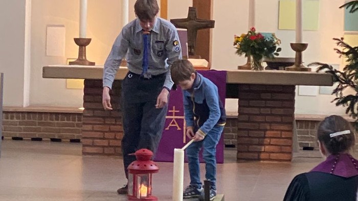 Zwei Jungen zünden eine Kerze in einer Kirche an, im Hintergrund sind ein Altar und Blumen zu sehen.