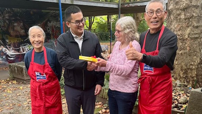 Vier Personen in Schürzen, eine hält einen gelben Gegenstand, posieren im Freien.