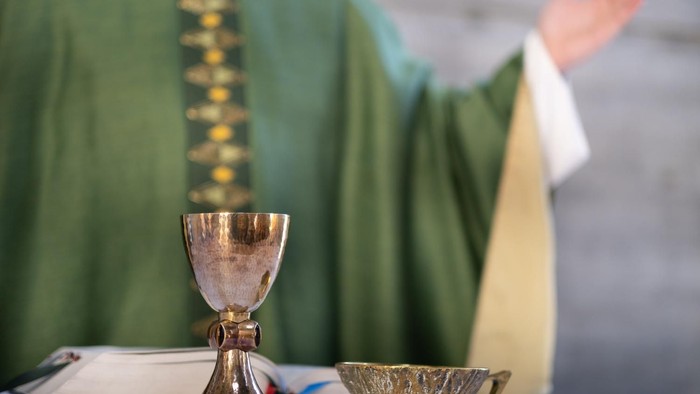 Ein Priester in grünen Messgewändern hält einen Kelch und eine Patene auf einem Altar.