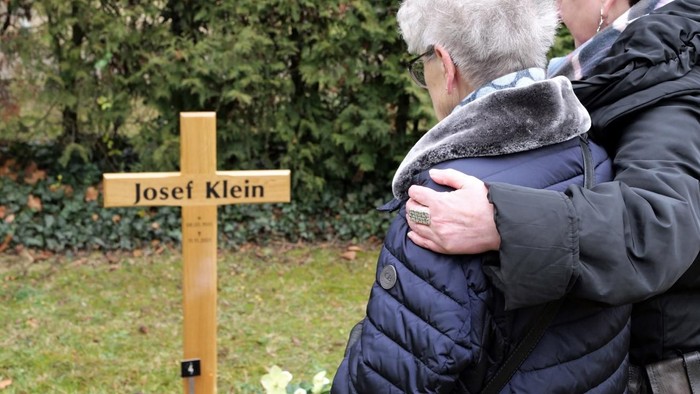Zwei Personen umarmen sich in einem Garten neben einem hölzernen Schild mit der Aufschrift *„Josef Klein“*.