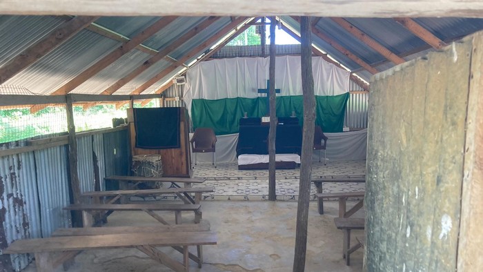 Ein überdachter Außenbereich mit Holzbänken und Tischen, grünen Vorhängen und einer rustikalen Atmosphäre