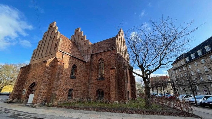 Gamle teglsteinskirke ved vej med træer og biler.
