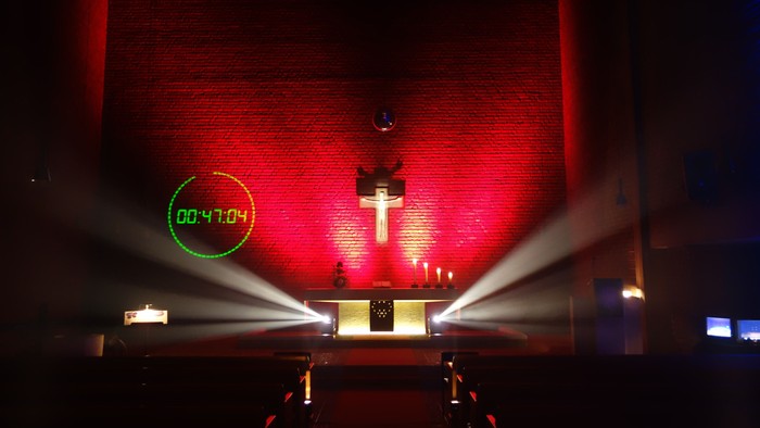 Auf dem Bild ist ein Altar mit darüber hängendem Kreuz zu sehen. Die Rückwand des Raumes leuchtet rot. Vom Altar selbst gehen weiße Lichtstrahlen aus, die in der Länge breiter werden. Ansonsten ist Kirchraum, in dem auch Bänke ein Reihen stehen, dunkel.