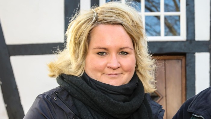 Eine Frau mit blondem Haar und schwarzer Schals steht vor einem Gebäude mit schwarzem und weißem Holz.