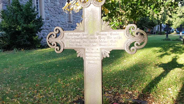 Ein Steinkreuz mit aufwendigen Verzierungen steht in einem grasbewachsenen Bereich mit Bäumen und einem steinernen Gebäude im Hintergrund.