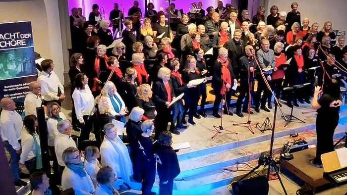 Zwei verschiedene Chöre, einer in weißer, einer in schwarzer Kleidung mit roten Tüchern, singt in einer Kirche, gesehen von oben links