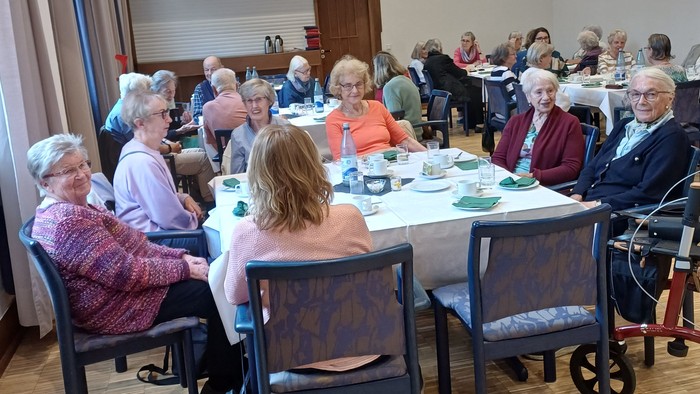 Gruppe älterer Menschen, die in einem gut beleuchteten Raum um Tische versammelt sind, einige sitzen im Rollstuhl.