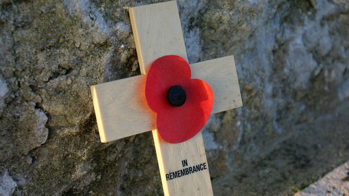 Wooden cross with red poppy against stone wall.