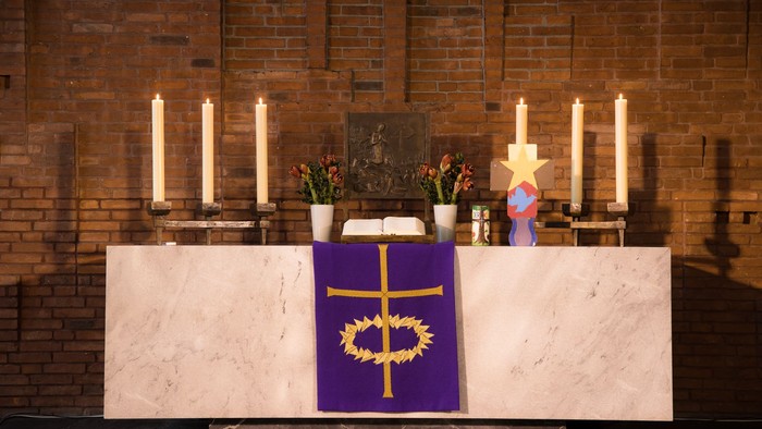 Altar mit violettem Tuch, goldenem Kreuz, Kerzen und Blumen.