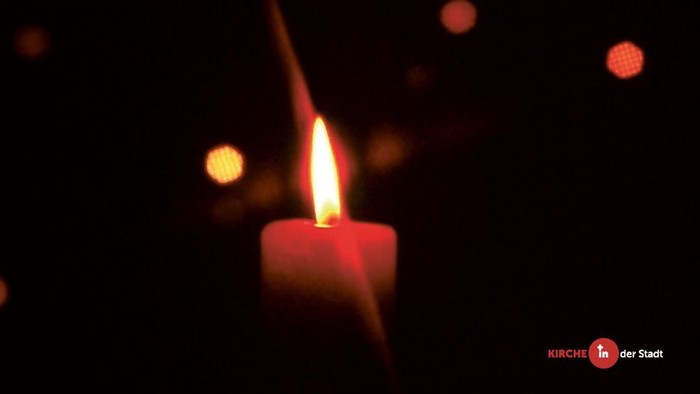 Ein brennendes Kerzenlicht vor dunklem Hintergrund mit Weihnachtsbeleuchtung und Text