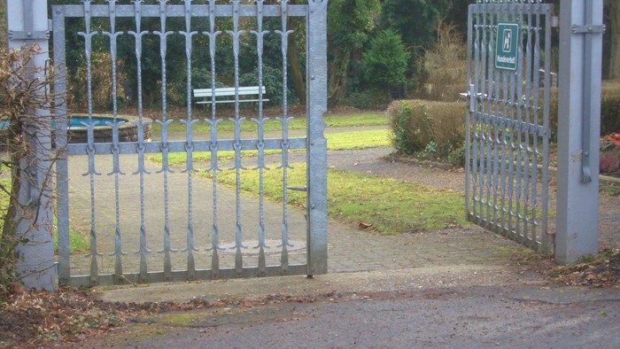 Das Bild zeigt ein offenes, weißes Tor mit spitzen Zäunen, das zu einem Park führt.