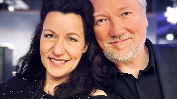 Ein Paar lächelt in die Kamera, die Frau trägt ein schwarzes Kleid, der Mann einen dunklen Anzug