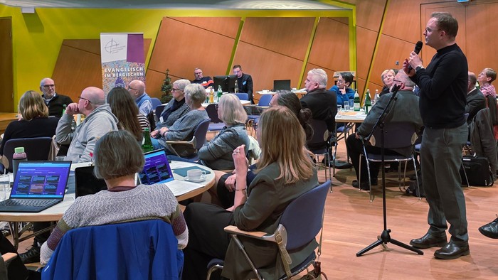 Blick in einen Tagungssaal mit vielen Menschen, die ihren Blick nach vorn auf eine Präsentation richten. Stehend ein Mann am Mikrofon, der spricht.