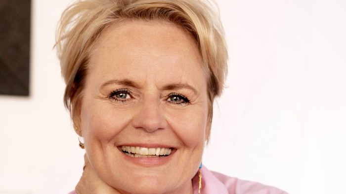 En kvinde med kort blondt hår, iført en lyserød skjorte og et grønt armbånd, smiler varmt.