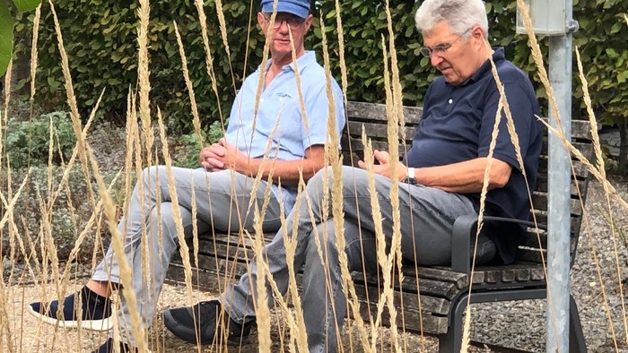 2 Männer sitzen mit überinandergeschlagenen Beinen im Gespräch vertieft auf einer Bank. Vor ihnen ausgebleichte Gräser, durch die der Blick des Betrachtenden geht.
