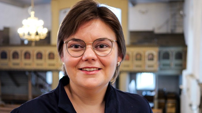 Eine Frau mit Brille und kurzen braunen Haaren lächelt in die Kamera. Im Hintergrund eine Kirchenempore