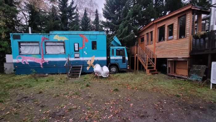 Ein blauer Bus mit bunten Mustern ist in der Nähe eines Holzhauses mit Hühnern in der Umgebung geparkt.