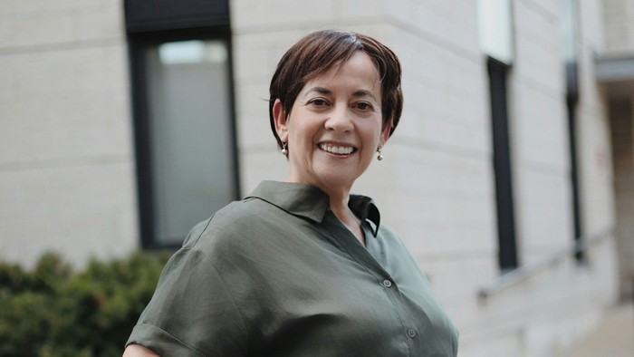 Frau in grünem Hemd und weißen Hosen, die draußen lächelt.