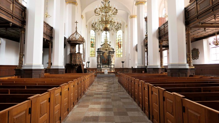Kirkeinteriør med træbænke, krystallysekrone og glasmosaikvinduer.