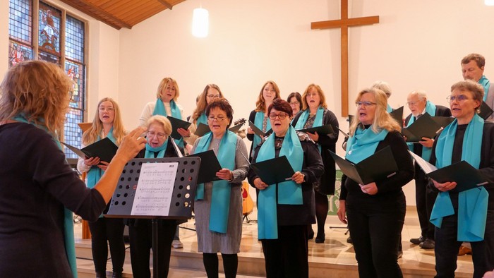 Ein Chor mit türkisblauen Schals singt in einer hellen Kirche. Im Hintergrund ein hölzernes Kreuz