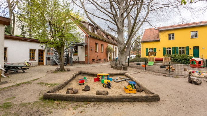Im Innenhof eines Kindergartens befindet sich ein Sandkasten mit bunten Spielzeugen und umgebenden Gebäuden.