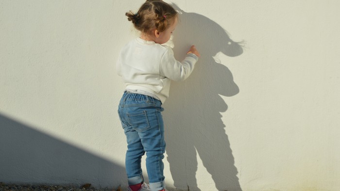 Ein junges Mädchen in einem weißen Hemd und blauen Jeans steht vor einer weißen Wand, ihr Schatten ist an der Wand sichtbar.