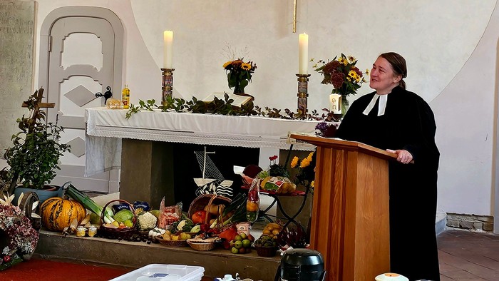 Ein Priester steht auf einer Kanzel in einer Kirche und hält eine Predigt. Der Altar ist mit Blumen und Kerzen geschmückt.