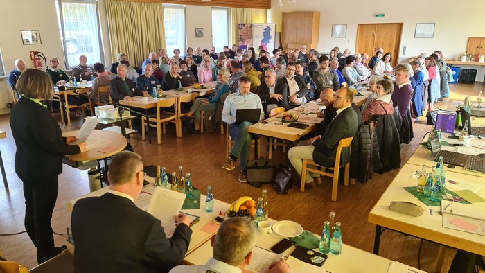 Große Gruppe von Menschen sitzt in einem Raum mit Tischen und Stühlen, einige stehen und unterhalten sich.