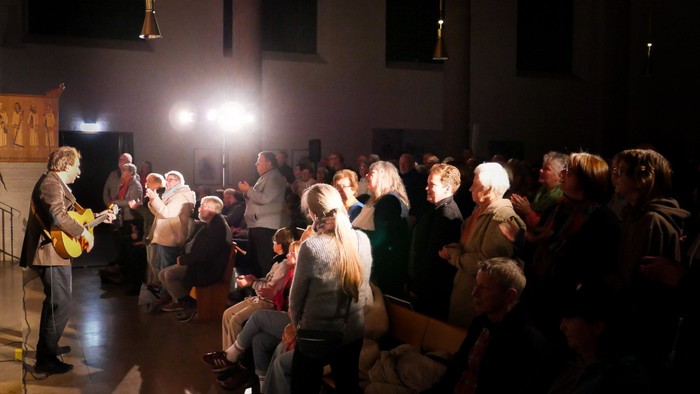 Ein Mann spielt Gitarre vor einem stehenden und klatschenden Publikum in einer Kirche