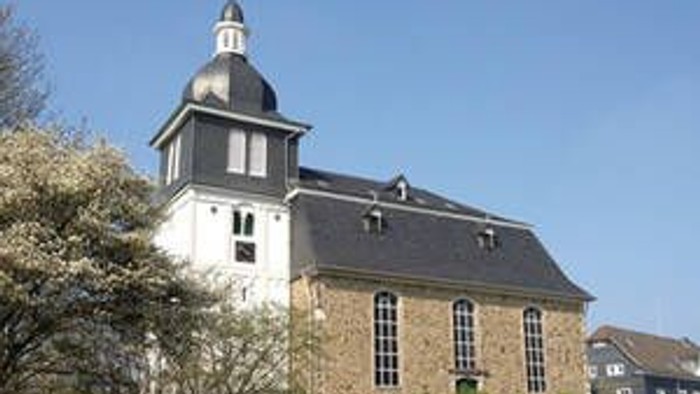 Das Bild zeigt eine Kirche mit einem Turm und einem grauen Dach, umgeben von Bäumen und Büschen.