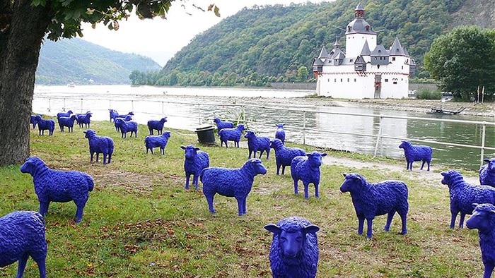 Blaue Schafskulpturen stehen auf Wiese vor Fluss und Schloss.