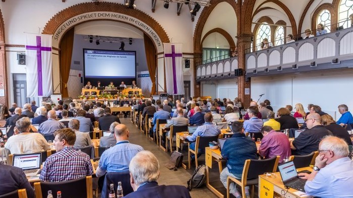 Großer Konferenzraum mit Personen, die an Tischen sitzen und einer Bühne mit Leinwand und Lautsprechern zugewandt sind.