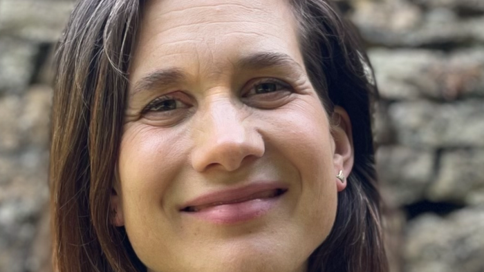 Eine Frau mit schulterlangem braunem Haar lächelt vor einer Steinmauer.