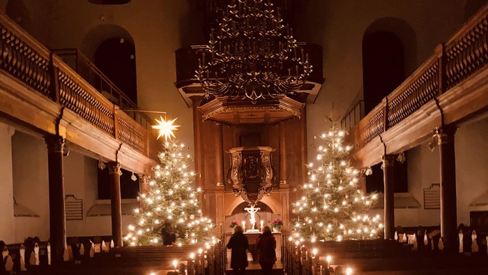 In der Kirche stehen zwei Weihnachtsbäume mit Lichtern, umgeben von Kirchenbänken und Kerzen.